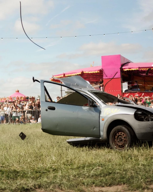 Damaged car with open door sits on grass with crowd and pink structure in the background under sunny sky, auto wreckage.