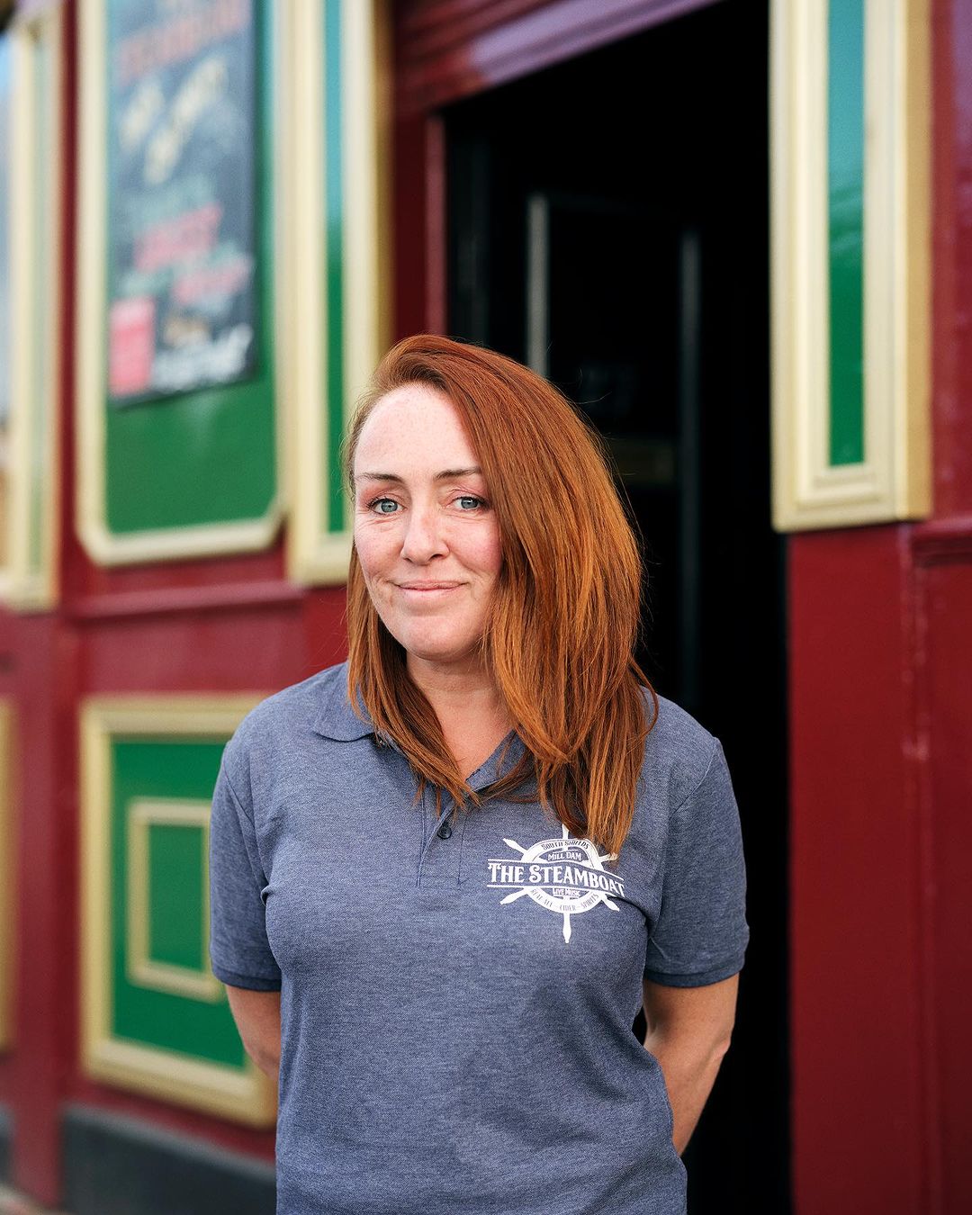 Portrait of a red-haired woman wearing a gray polo shirt with a logo, standing outside a building with red and green trim.