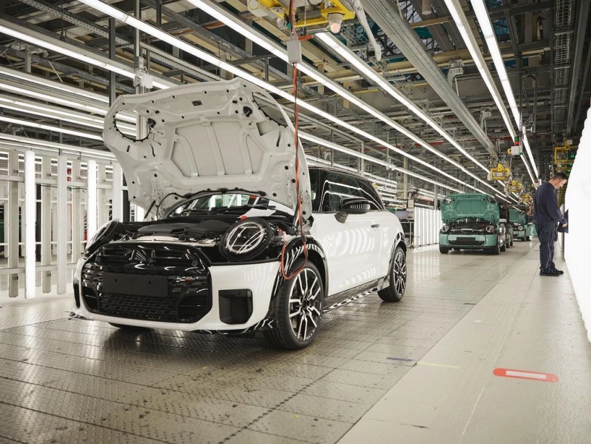 Automotive factory line showcasing a white Mini Cooper with its hood open, surrounded by manufacturing equipment and factory workers.