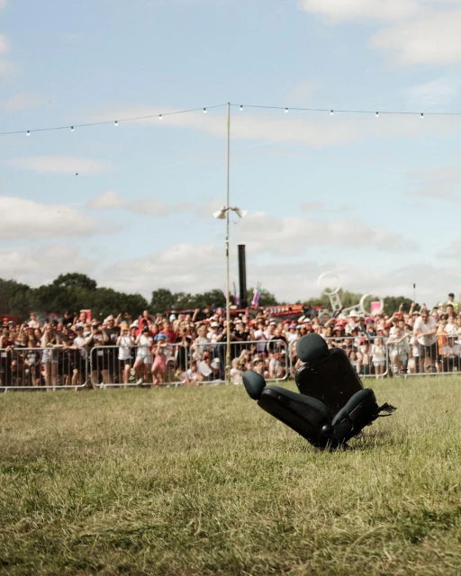 Black car seat sits on green grass at an outdoor gathering with a crowd of people and white metal barriers in the background.