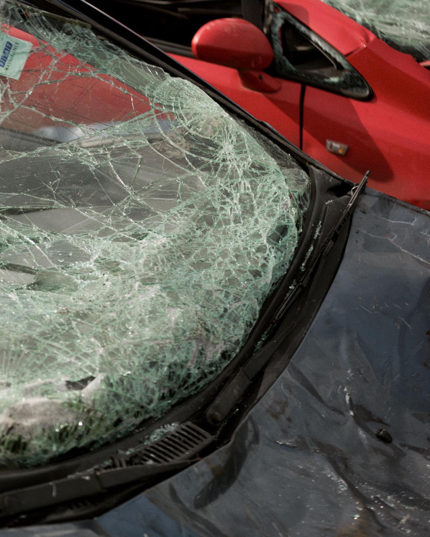 Close-up of a car's shattered windshield with a web-like crack pattern, revealing parts of another red car in the blurred background.