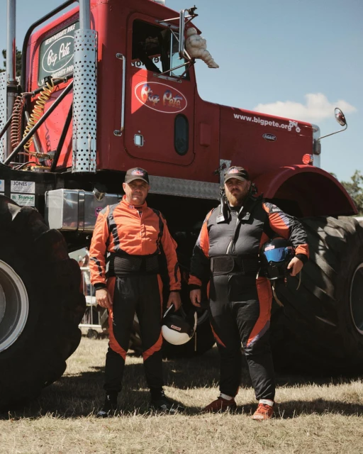 Two adult males stand in front of a large monster truck with oversized tires, wearing matching jumpsuits and holding helmets.