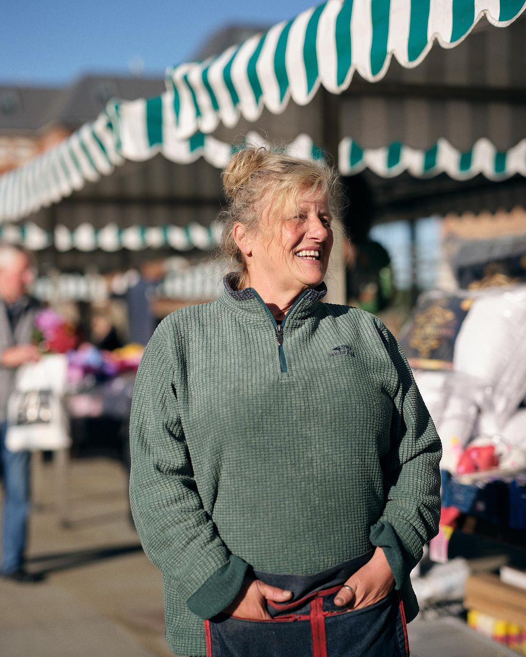 Smiling woman at an outdoor market, wearing a green fleece and denim apron with red straps, with striped canopy tents visible.