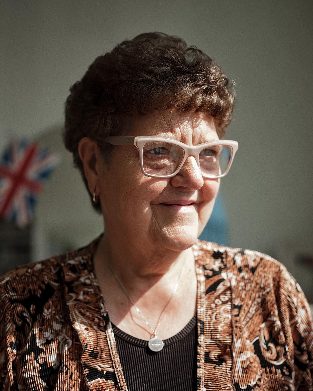 Elderly woman with short brown hair and glasses, wearing a patterned top and necklace, smiling slightly with a Union Jack in the background.