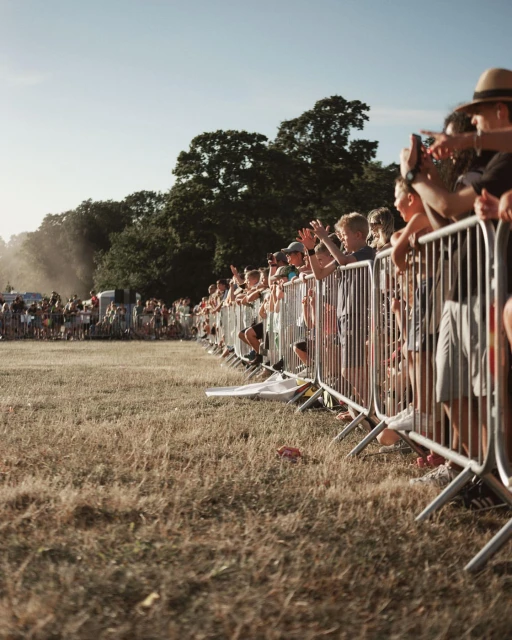 Crowd of people gathers behind a metal fence in an open field with trees in the background on a sunny day. People are standing and raising their arms.