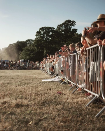 Crowd of people gathers behind a metal fence in an open field with trees in the background on a sunny day. People are standing and raising their arms.