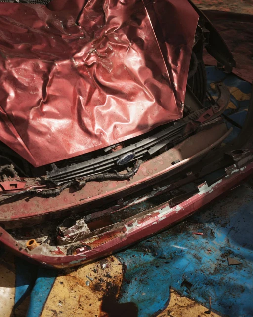 Close-up of a wrecked, red car with a crumpled hood and damaged bumper, suggesting a vehicle accident with significant damage.