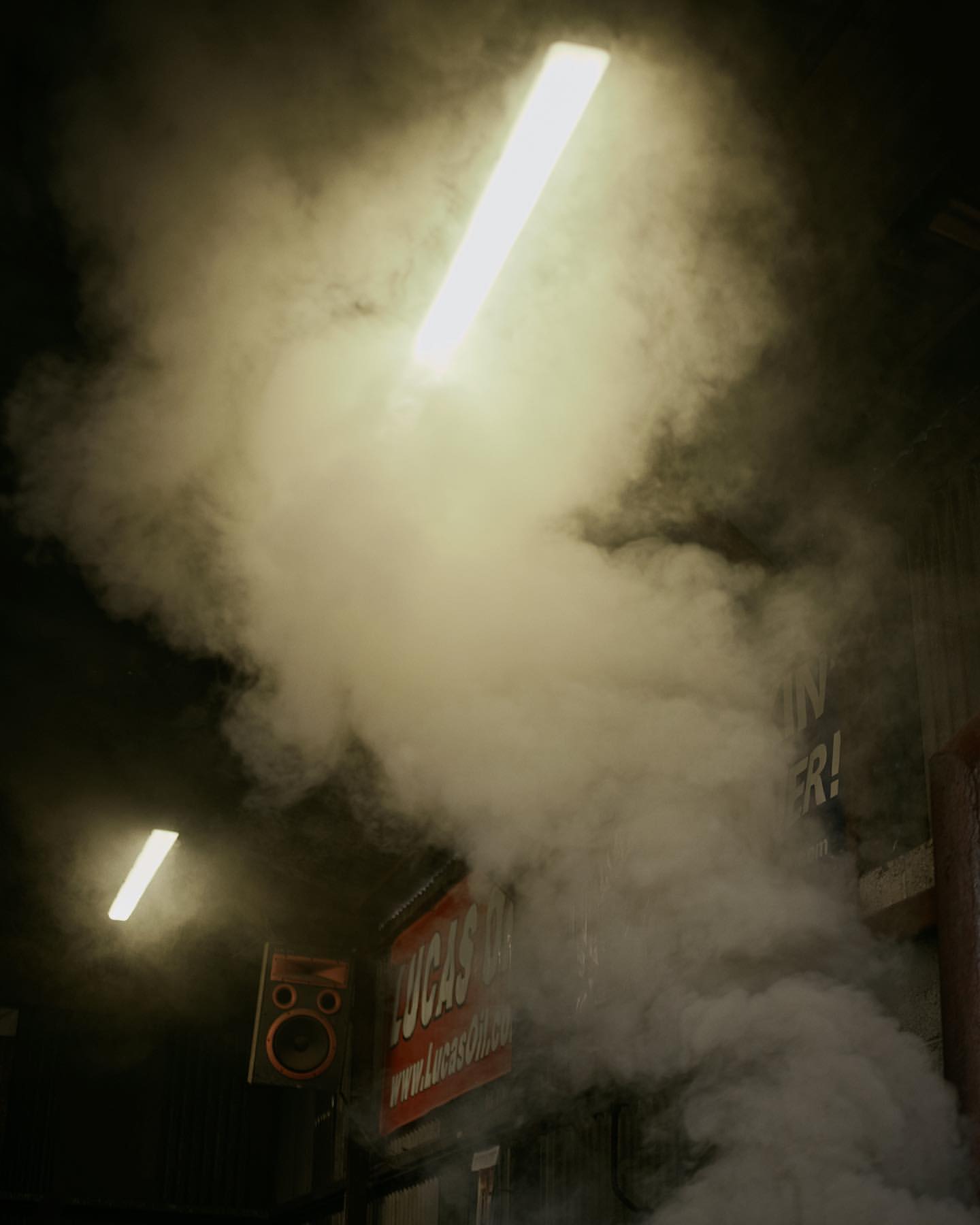 Dense smoke and vapor fills an indoor space, illuminated by bright fluorescent lights. A sign is partially visible behind the smoke.