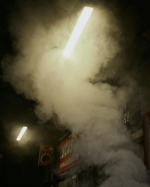 Dense smoke and vapor fills an indoor space, illuminated by bright fluorescent lights. A sign is partially visible behind the smoke.
