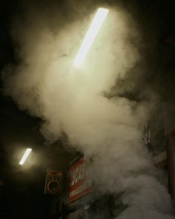 Dense smoke and vapor fills an indoor space, illuminated by bright fluorescent lights. A sign is partially visible behind the smoke.