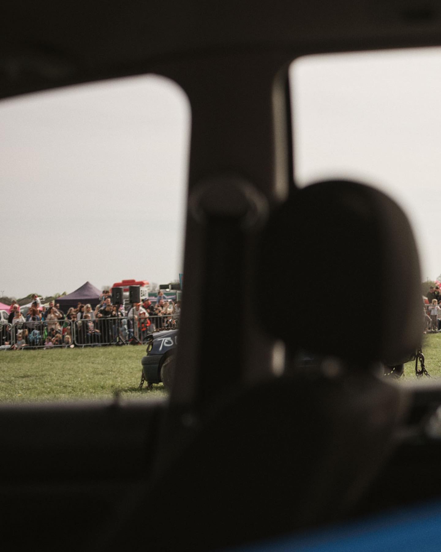 View through a car window of an outdoor event with a crowd of people gathered on a grassy field, tents, and a small vehicle.