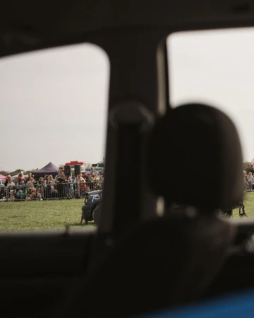 View through a car window of an outdoor event with a crowd of people gathered on a grassy field, tents, and a small vehicle.