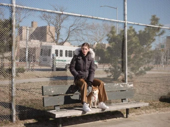 A woman in a dark hooded puffer jacket and brown leather trousers seated on a park bench with a Shiba Inu dog in London