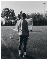 A soccer player standing on a pitch facing a goal net, captured in a monochrome vertical composition.