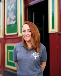 A woman with red hair wearing a grey polo shirt standing outside a dark doorway with red and green framing.