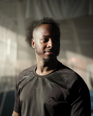 Portrait of a black man wearing a dark t-shirt with geometric chest patterning, captured in soft indoor lighting.