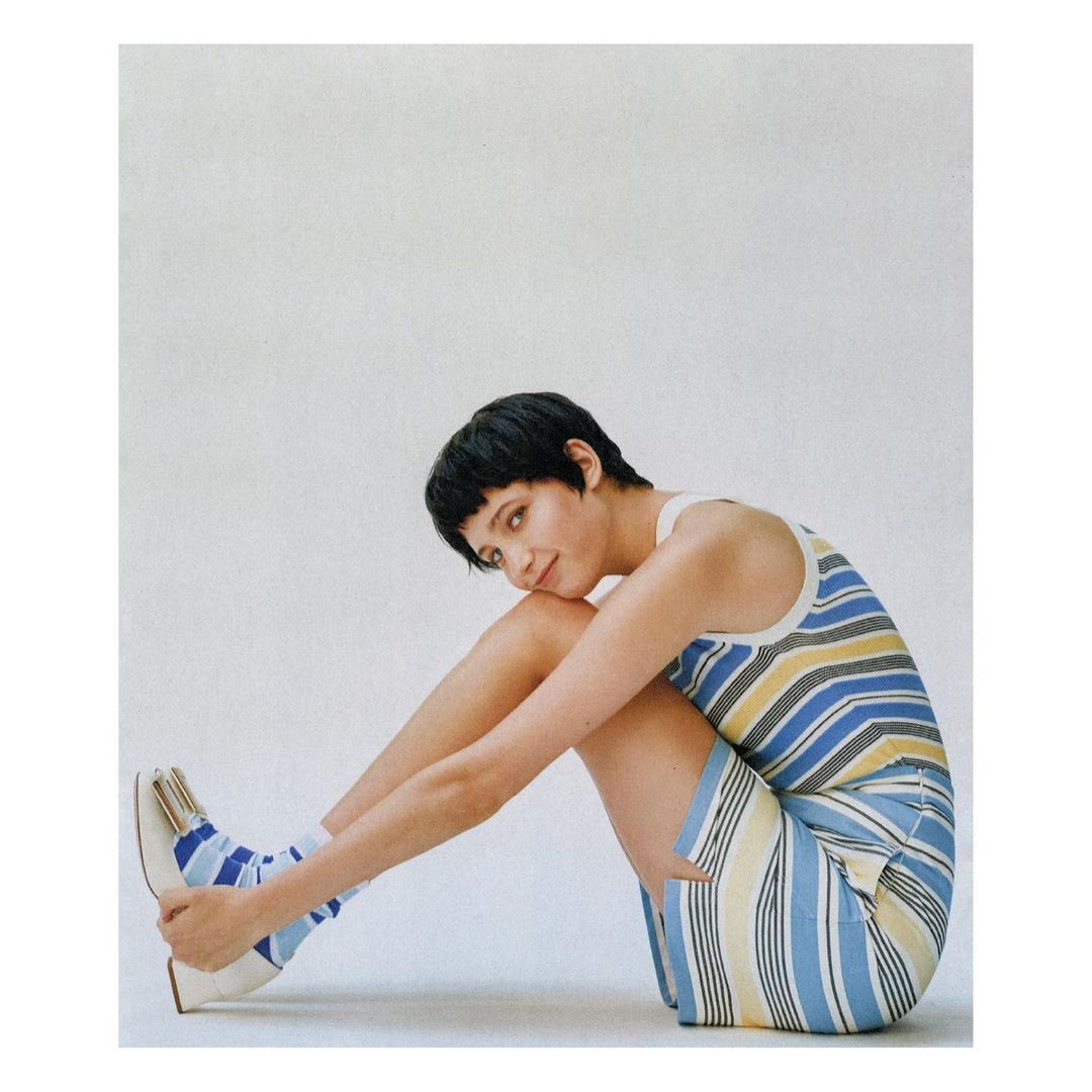 A young woman in a striped dress and striped socks, sitting with legs crossed against a white background in a fashion studio portrait.