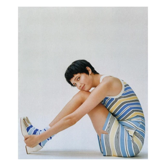 A young woman in a striped dress and striped socks, sitting with legs crossed against a white background in a fashion studio portrait.