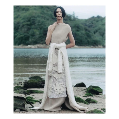 Woman with short black hair wearing a long cream knit dress standing on a beach with mossy rocks by the water.