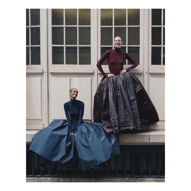 Two women in elegant dresses pose in front of a window. One woman is sitting, the other standing in a burgundy dress. Fashion shoot.