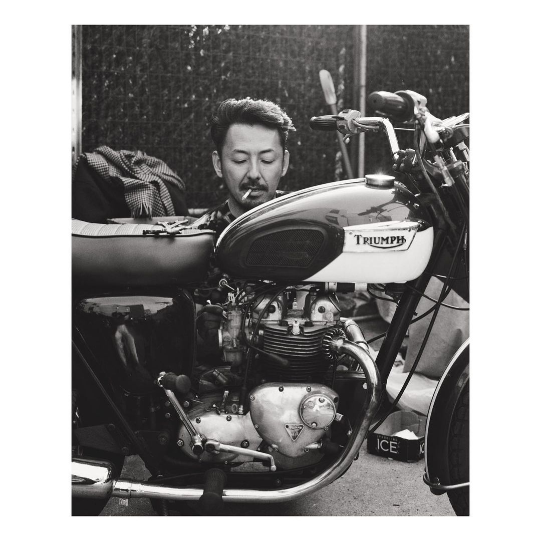 Monochrome shot of a stylish man with a cigarette, posing on a vintage Triumph motorcycle, with a blurred urban backdrop.