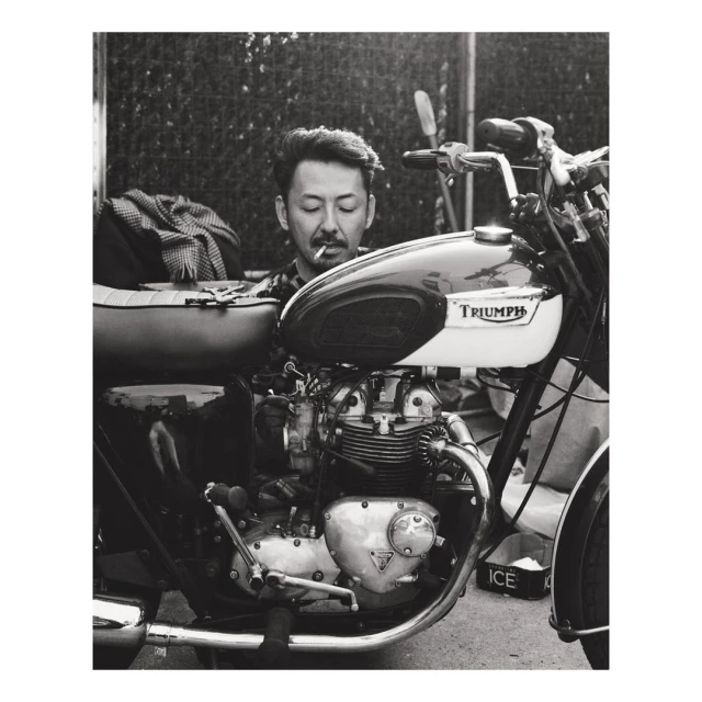 Monochrome shot of a stylish man with a cigarette, posing on a vintage Triumph motorcycle, with a blurred urban backdrop.