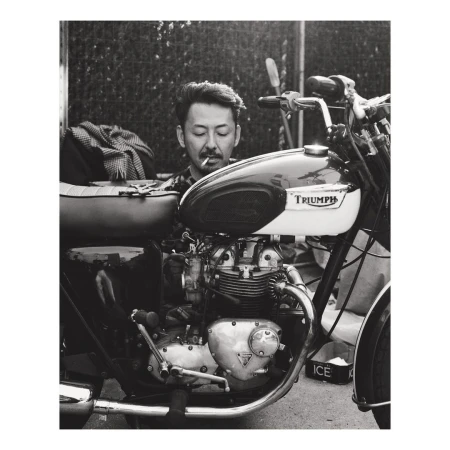 Monochrome shot of a stylish man with a cigarette, posing on a vintage Triumph motorcycle, with a blurred urban backdrop.