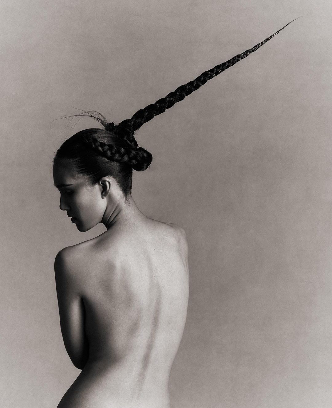 Monochrome portrait of a nude woman from the back, her head turned to the left, featuring an elaborately braided hairstyle extending upwards.