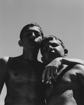 Black-and-white photo of two young, shirtless boys embracing outdoors under a clear sky. Low angle capturing their upper bodies.