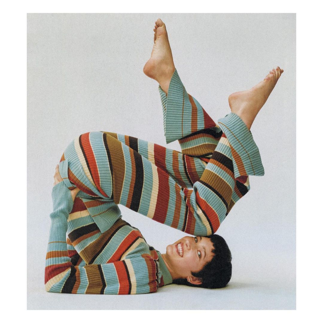 A woman in a multicolor striped knit outfit is lying upside down in a yoga position against a white backdrop and looking at the camera.