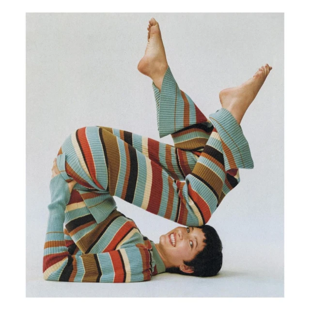 A woman in a multicolor striped knit outfit is lying upside down in a yoga position against a white backdrop and looking at the camera.