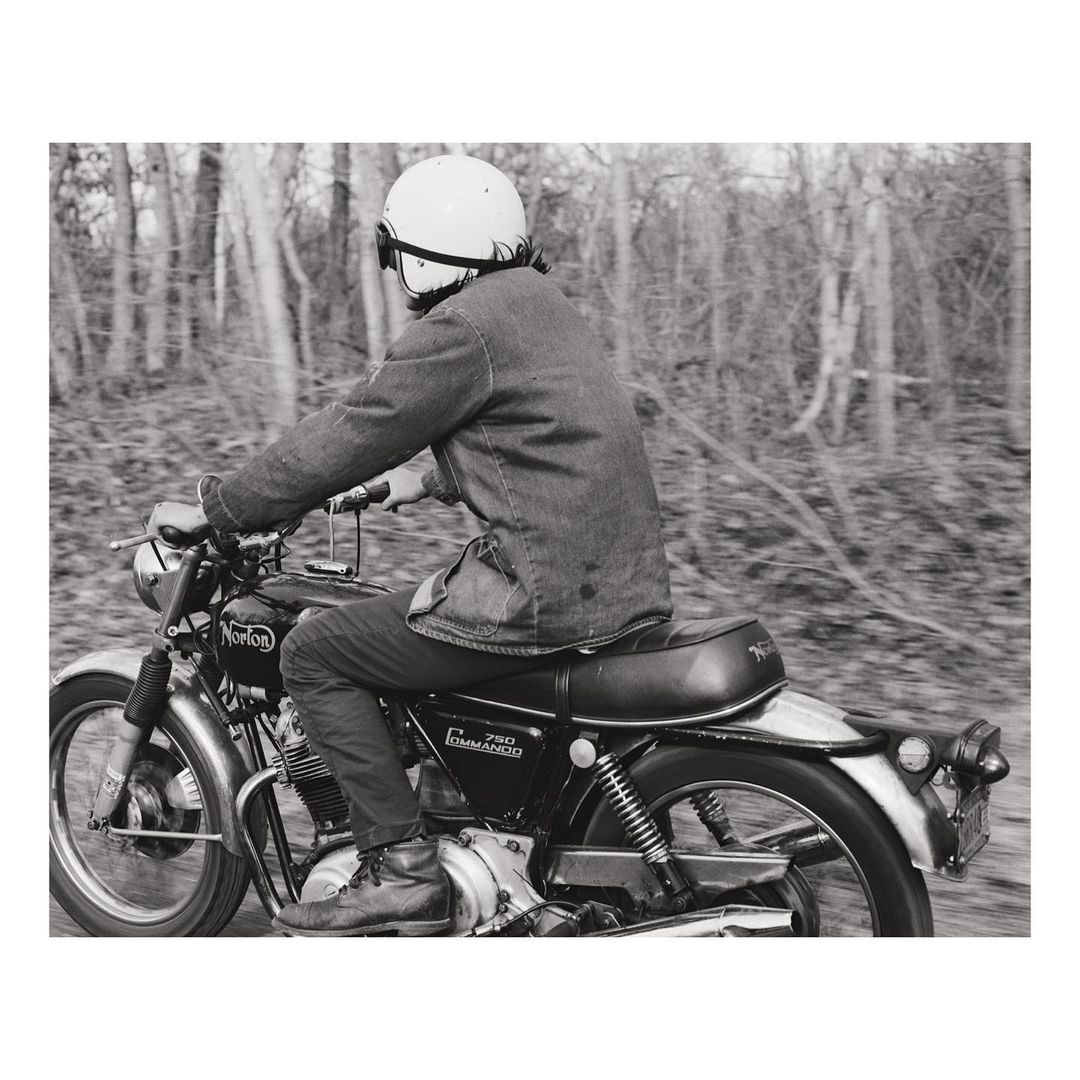 Vintage motorcycle rider with helmet navigates a wooded road in a monochromatic photo, capturing a sense of speed and classic style.