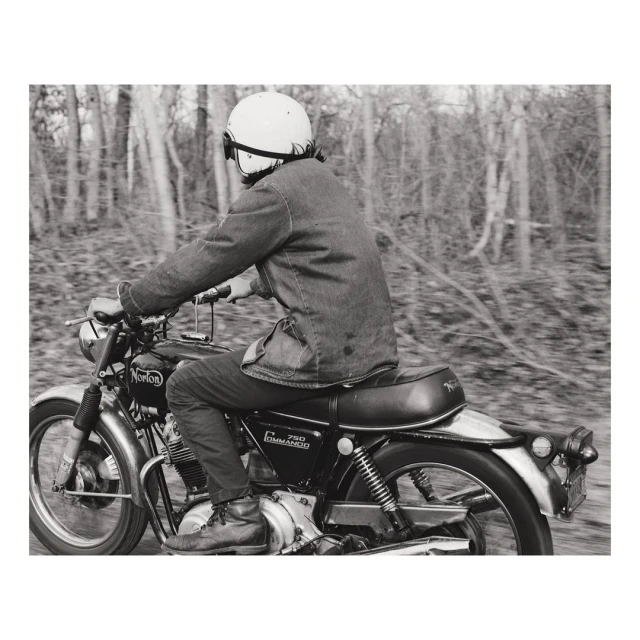 Vintage motorcycle rider with helmet navigates a wooded road in a monochromatic photo, capturing a sense of speed and classic style.