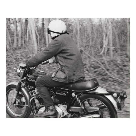 Vintage motorcycle rider with helmet navigates a wooded road in a monochromatic photo, capturing a sense of speed and classic style.