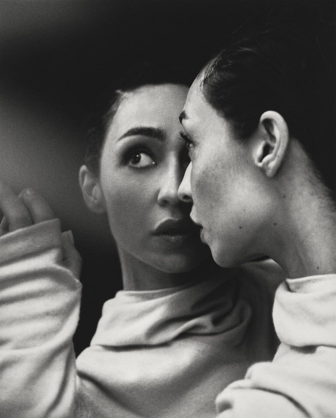 Monochrome portrait of an adult woman gazing with focused expression, her reflection visible in a mirror indoors.