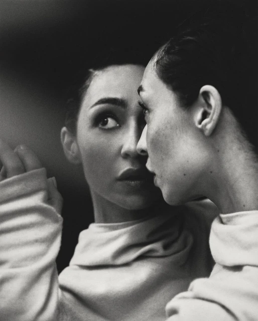 Monochrome portrait of an adult woman gazing with focused expression, her reflection visible in a mirror indoors.