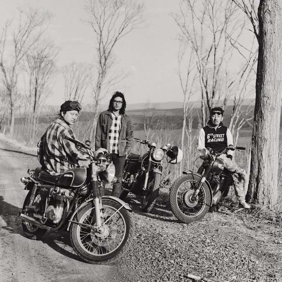 Three motorcyclists with their vintage motorcycles pose on a rural road in black and white, showcasing a moment of camaraderie.