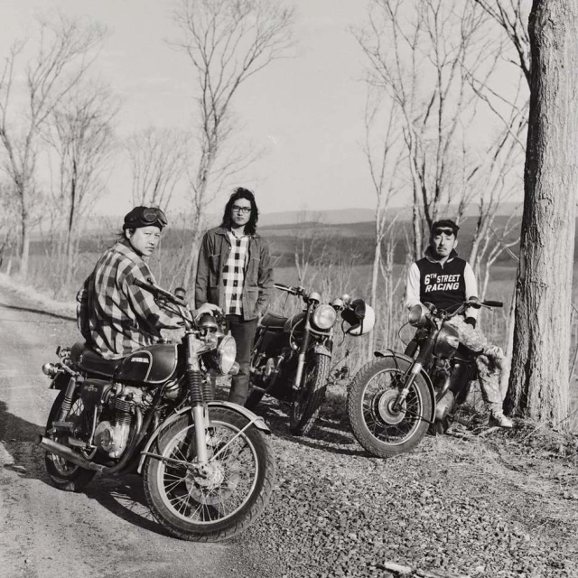 Three motorcyclists with their vintage motorcycles pose on a rural road in black and white, showcasing a moment of camaraderie.