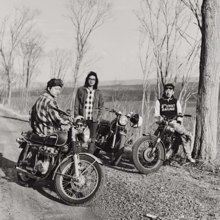 Three motorcyclists with their vintage motorcycles pose on a rural road in black and white, showcasing a moment of camaraderie.