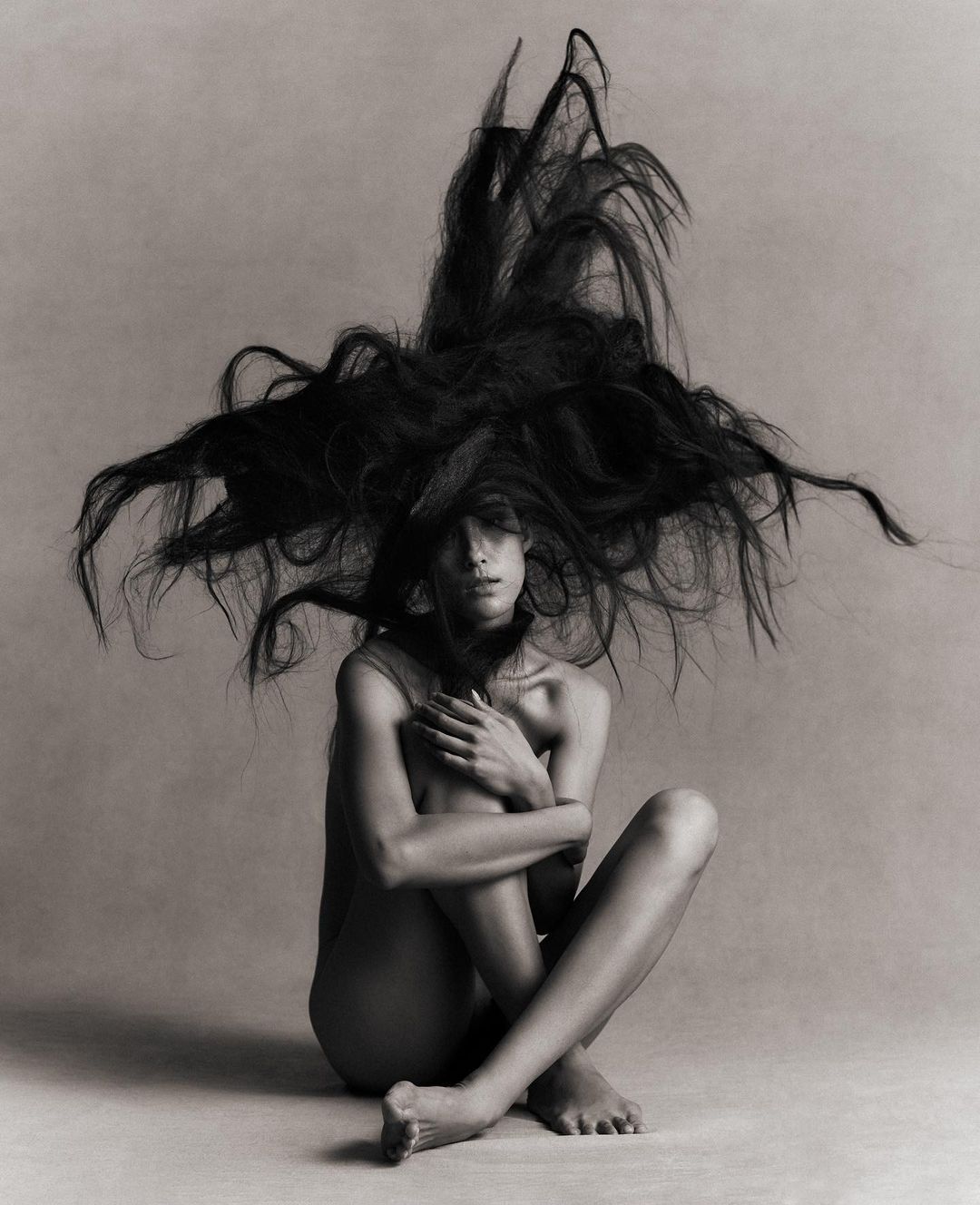 Artistic black and white portrait of a slender woman with long hair explosively surrounding her, seated with arms around her legs.
