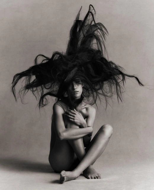 Artistic black and white portrait of a slender woman with long hair explosively surrounding her, seated with arms around her legs.