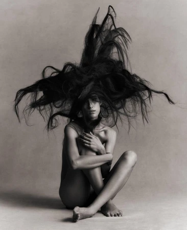 Artistic black and white portrait of a slender woman with long hair explosively surrounding her, seated with arms around her legs.