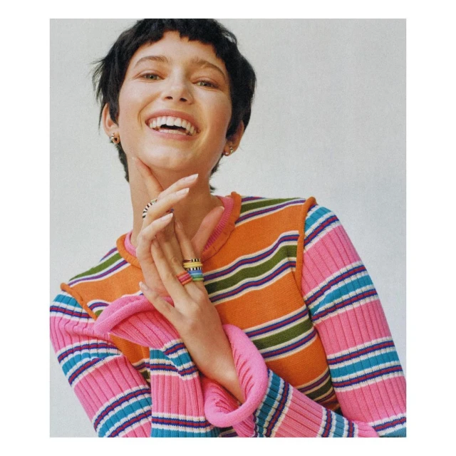 Smiling woman in a striped long-sleeve sweater with hands near her face, accessorized with multiple rings, in a light-filled setting.