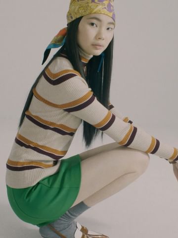 Asian woman wearing a patterned headscarf, striped turtleneck, and green skirt, squatting against a plain background.