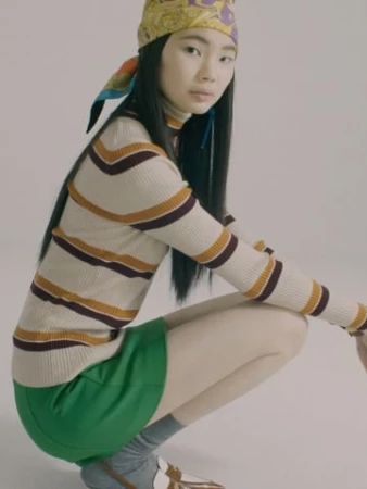 Asian woman wearing a patterned headscarf, striped turtleneck, and green skirt, squatting against a plain background.