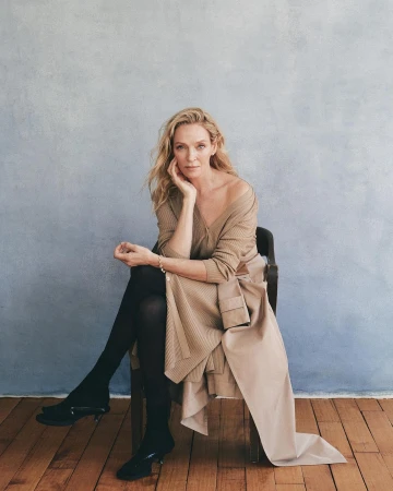 Blond woman in a tan sweater dress with black tights and heels sitting on a wooden chair in front of a textured blue wall.