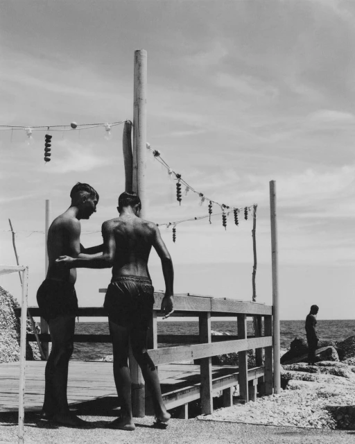 Black and white photo of two young men standing on a wooden platform by the sea, with a string of lights overhead and another man in the distance.