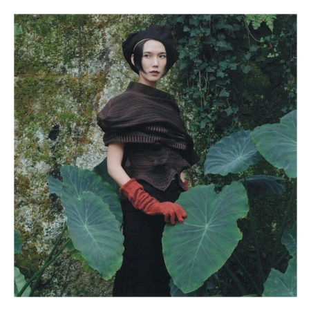 Woman in dark beret and brown knit top wearing orange gloves standing among giant green tropical leaves.