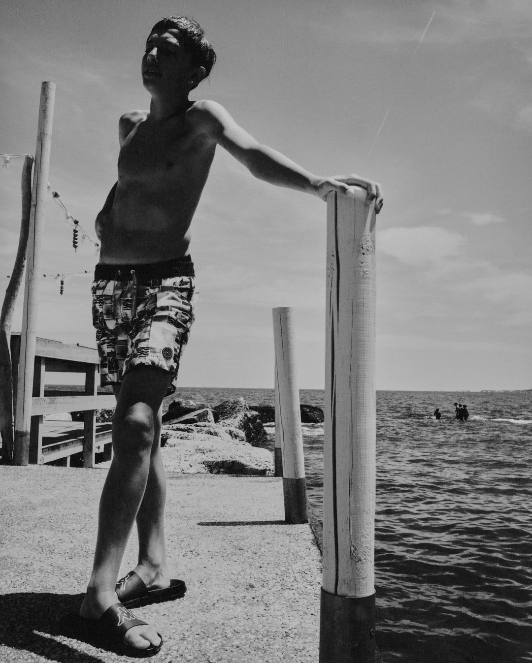 Black and white photo of a shirtless teenager wearing patterned shorts leaning on a wooden post next to the ocean on a sunny day.