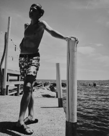 Black and white photo of a shirtless teenager wearing patterned shorts leaning on a wooden post next to the ocean on a sunny day.